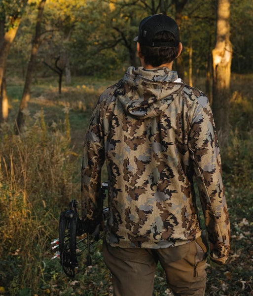 Person in outdoor clothing crouching in a wooded area.